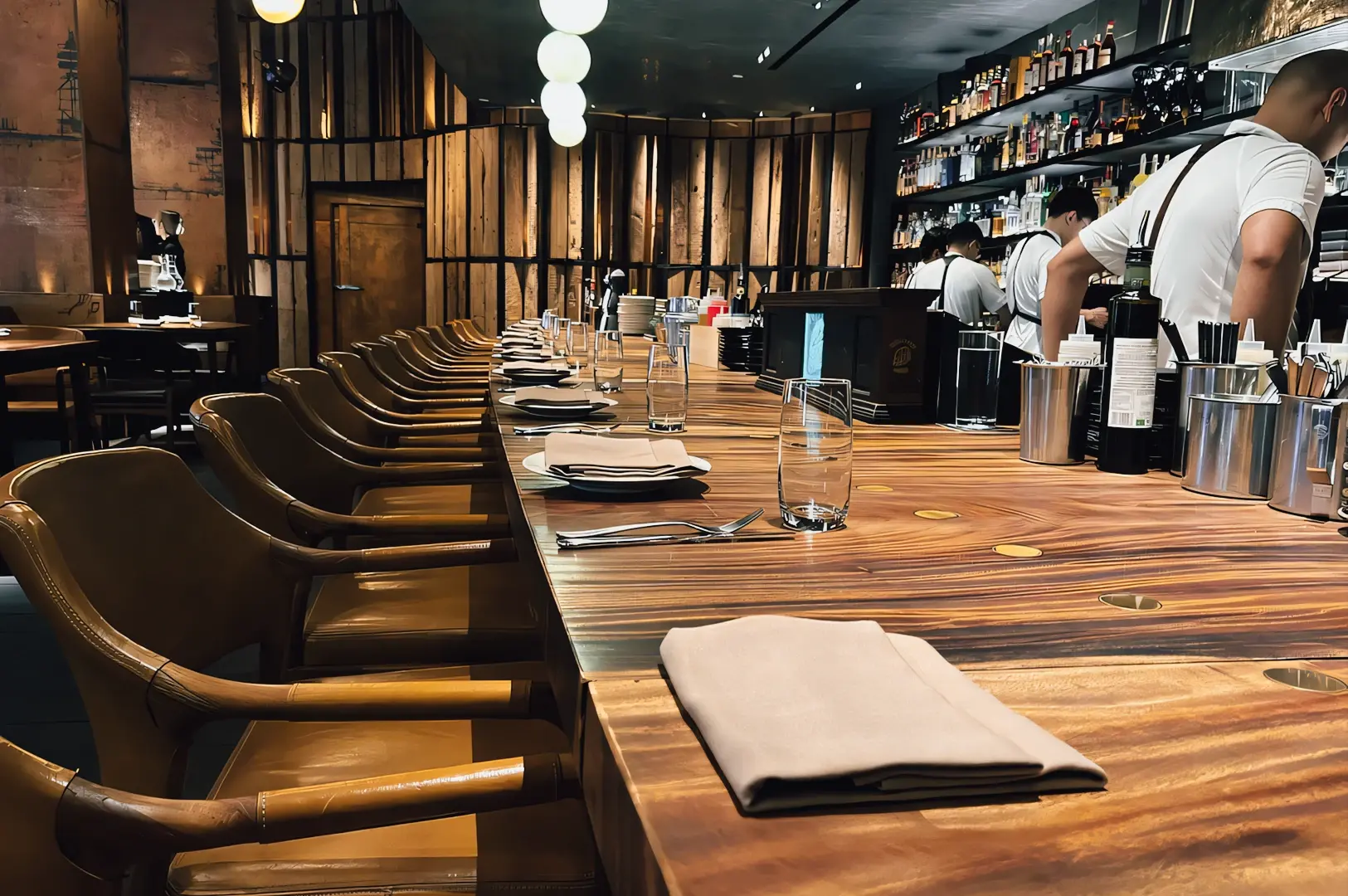 Contemporary restaurant bar with a long wooden counter, neatly set place settings, and open shelves of bottles, photographed from a low eye‑level linear angle highlighting chef’s counter dining and modern hospitality design.