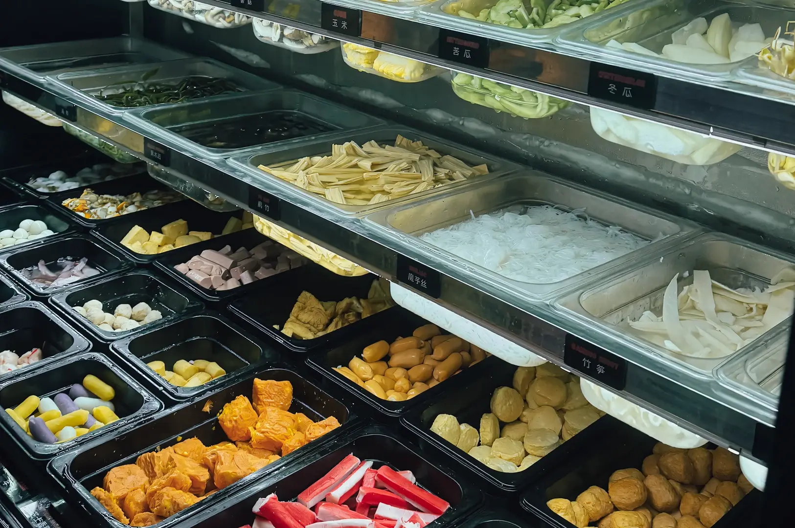  High-angle view of A Hot Hideout’s self-service mala ingredient bar at NEX Mall, showing neatly arranged trays of vegetables, fish cakes, tofu, mushrooms, and meats for DIY mala hotpot selection in Singapore.