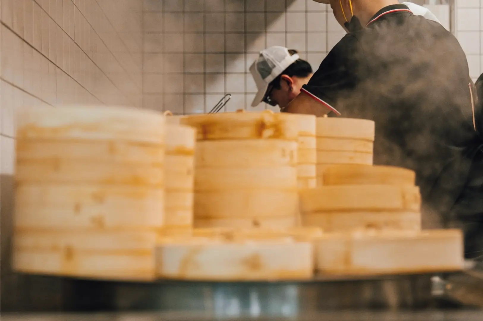 Low-angle, shallow depth-of-field shot of stacked bamboo steamers in the foreground with steam rising, while kitchen staff work in the background, capturing live dim sum preparation in a Hong Kong–style open kitchen.