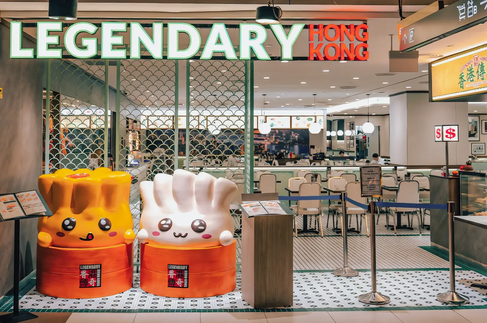 Wide-angle, eye-level shot of the entrance to Legendary Hong Kong restaurant, featuring bright illuminated signage, queue barriers, and two large cartoon-style dim sum mascots at the front, showcasing a modern Hong Kong–style dining concept inside a shopping mall.