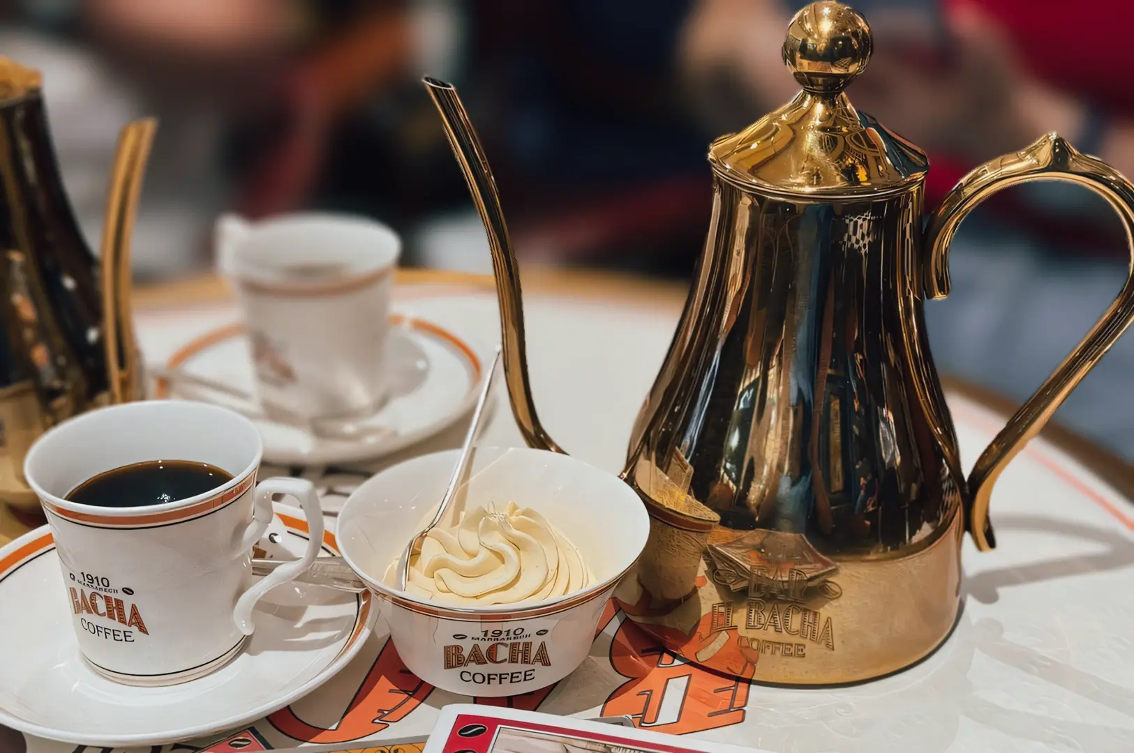 Close-up table-level shot of a traditional Bacha Coffee service with a gold coffee pot, espresso cups, and a bowl of whipped cream served on a marble café table.