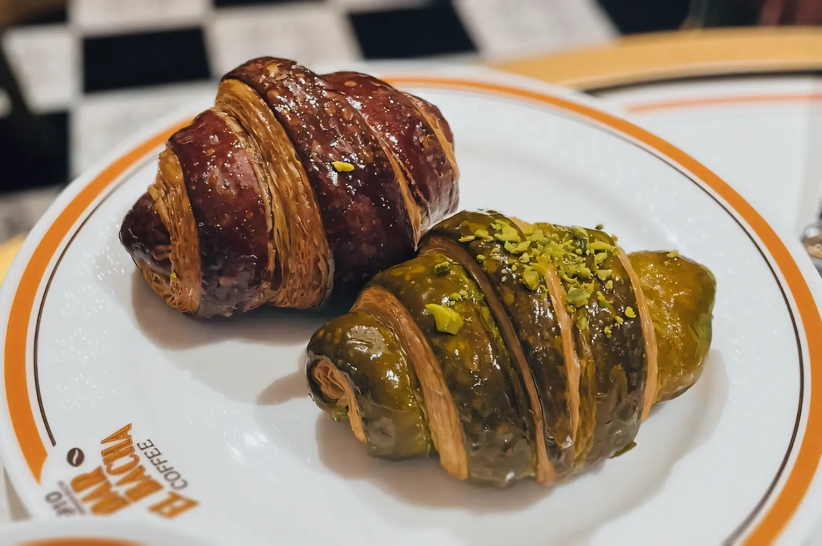 Overhead close-up of two glazed croissants on a branded plate, one chocolate and one pistachio, served at Bacha Coffee as part of a luxury café pastry selection.