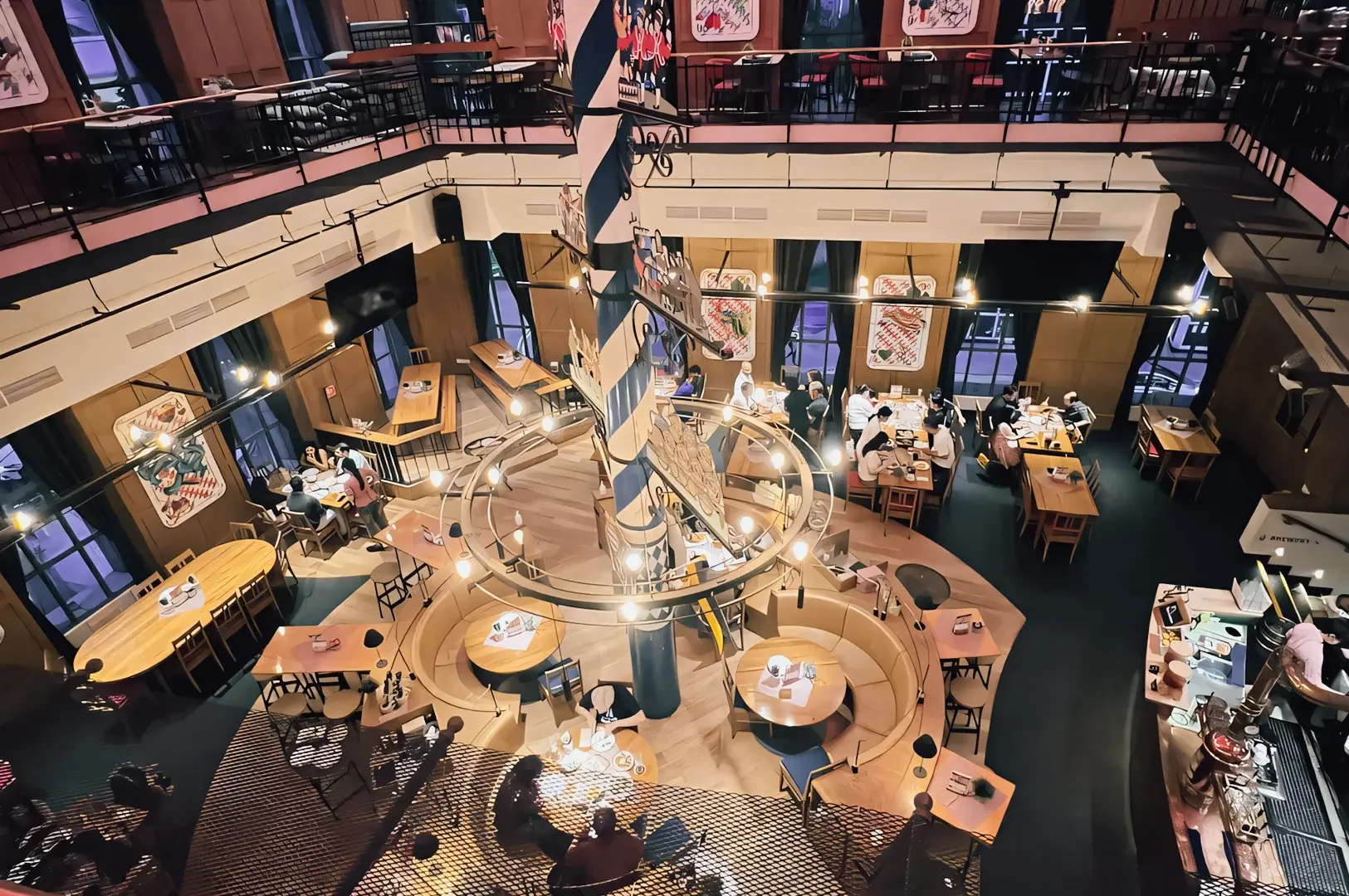 Multi‑level restaurant interior with communal tables, circular seating, and a central sculptural column, photographed from a high‑angle overhead view showcasing social dining, architectural lighting, and layered spatial design.