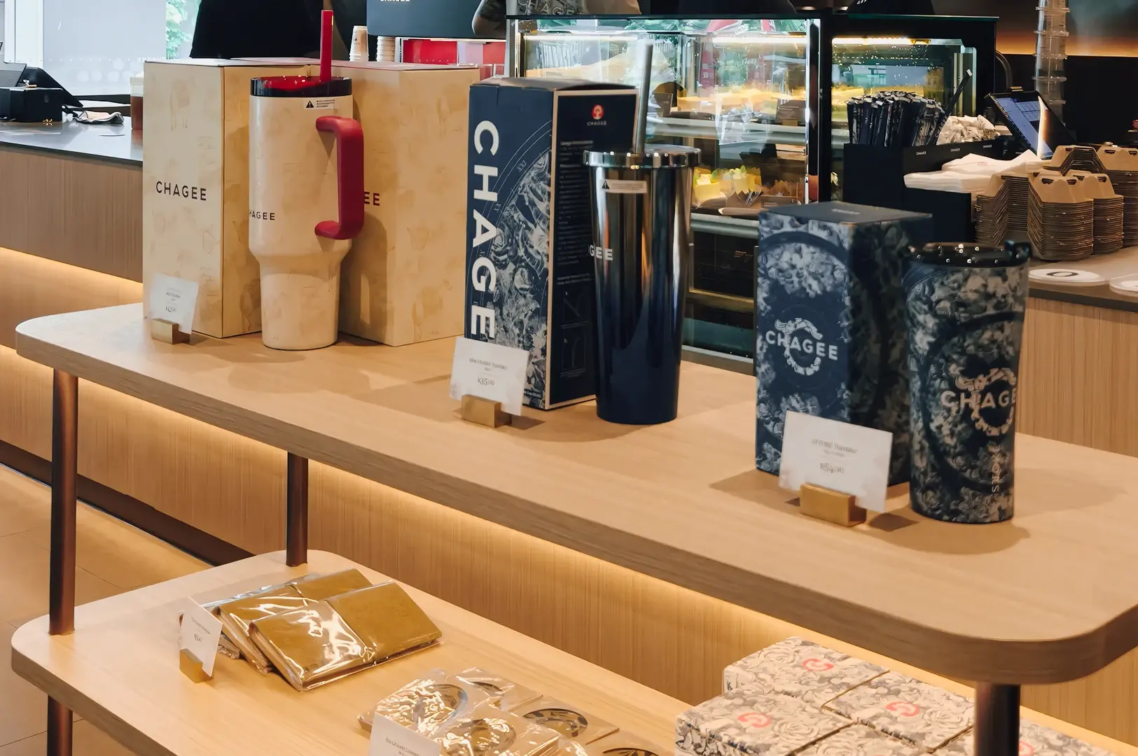 Eye‑level wide shot of a CHAGEE retail merchandise display at Causeway Point Singapore, showing branded tumblers, reusable cups, gift boxes, and tea accessories arranged on wooden shelves inside the tea café.