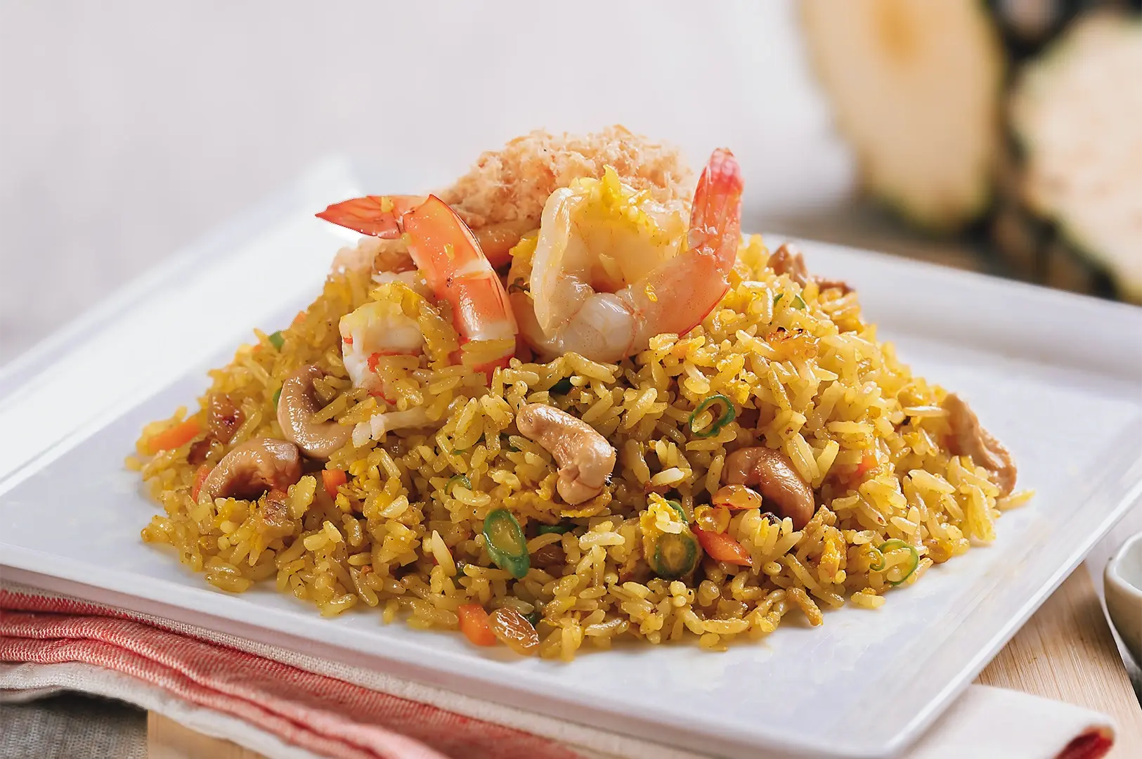 Close‑up food photograph taken from a slightly elevated angle of fried rice topped with prawns, cashews, and herbs, served on a white square plate with soft natural lighting.