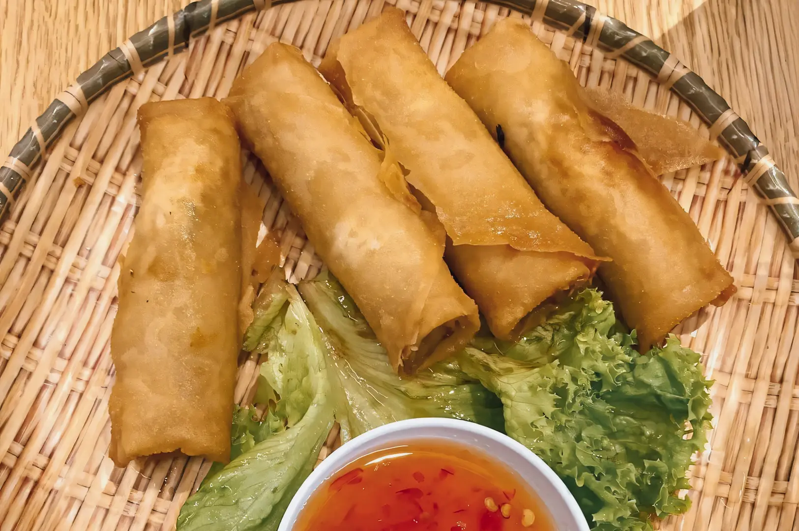 Top‑down close‑up shot of golden fried spring rolls arranged in a wicker basket with lettuce garnish and a bowl of sweet chili dipping sauce on a wooden surface.
