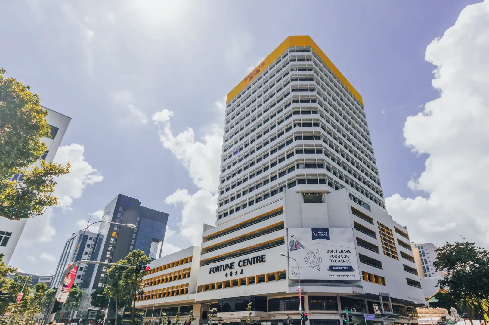 Low-angle, wide-angle exterior shot of Fortune Centre mall in Singapore, showing the multi-storey white building with yellow accents, the ‘Fortune Centre’ sign, surrounding city streets, traffic lights, trees, and a bright blue sky with scattered clouds in daylight.