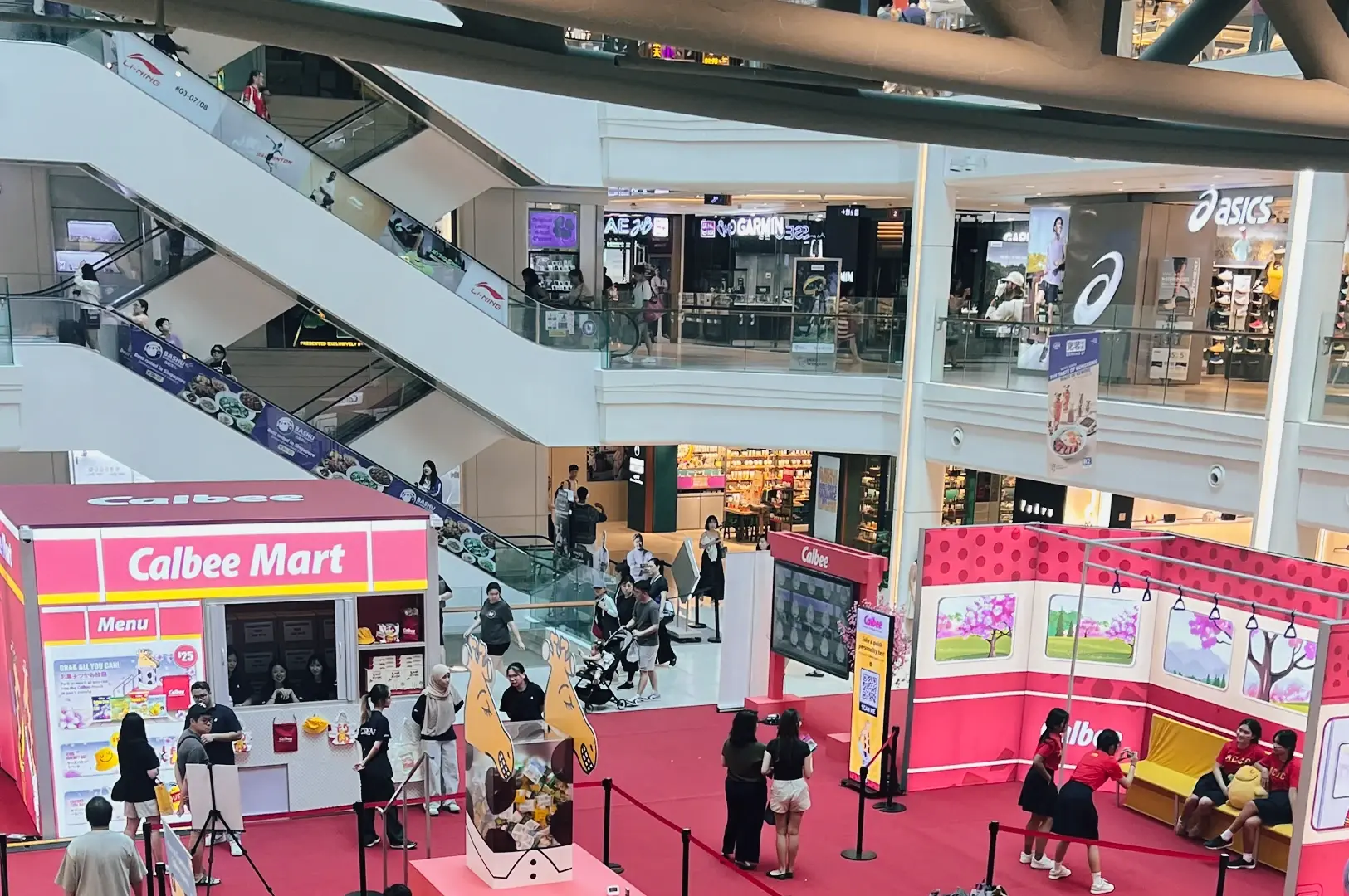 High-angle wide shot of Plaza Singapura interior atrium with escalators and Calbee pop-up event, highlighting vibrant retail activity and family-friendly shopping experience in Singapore.