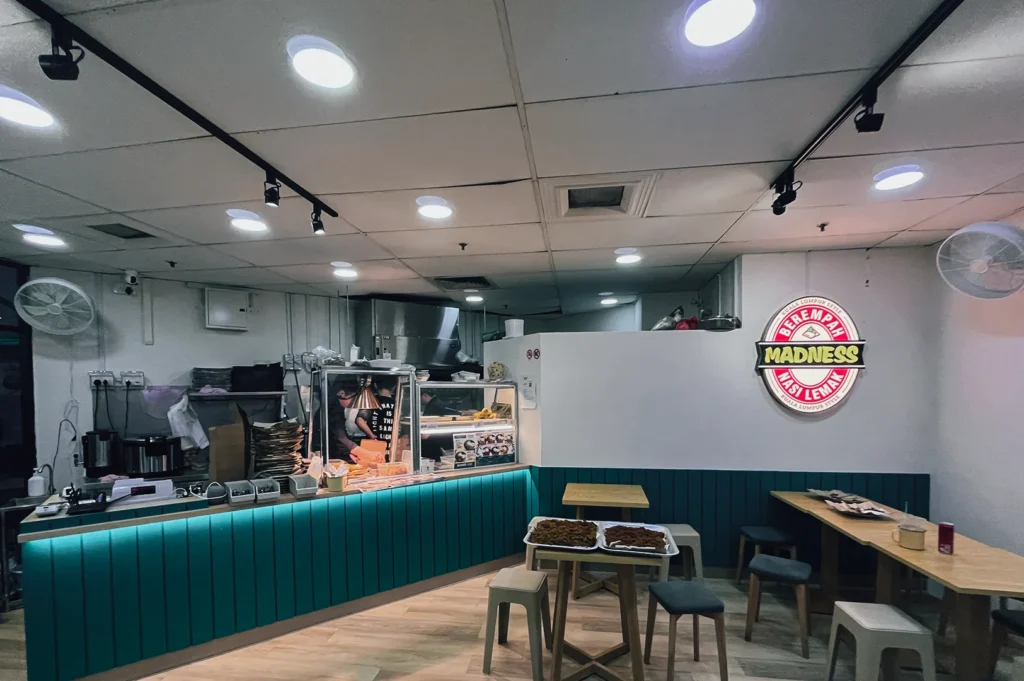 Wide-angle interior shot of Madness Nasi Lemak restaurant featuring open kitchen counter, modern casual dining tables, teal wall panels, and branded nasi lemak logo on the wall.