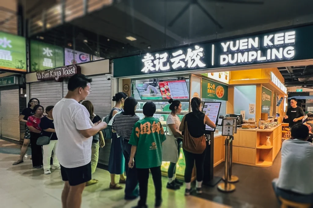 Eye-level candid shot of queue at Yuen Kee Dumpling with ordering kiosk and illuminated sign inside Fortune Centre food hub.