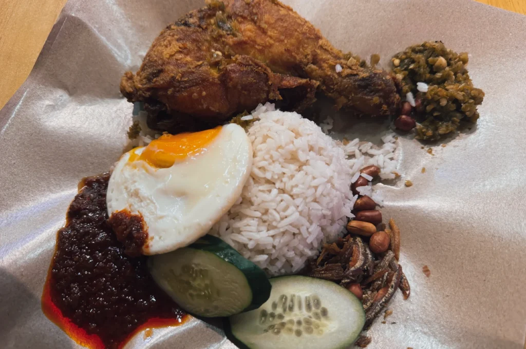 Close-up overhead shot of nasi lemak with crispy fried chicken, coconut rice, sambal, soft-boiled egg, anchovies, peanuts, and cucumber served on brown paper.