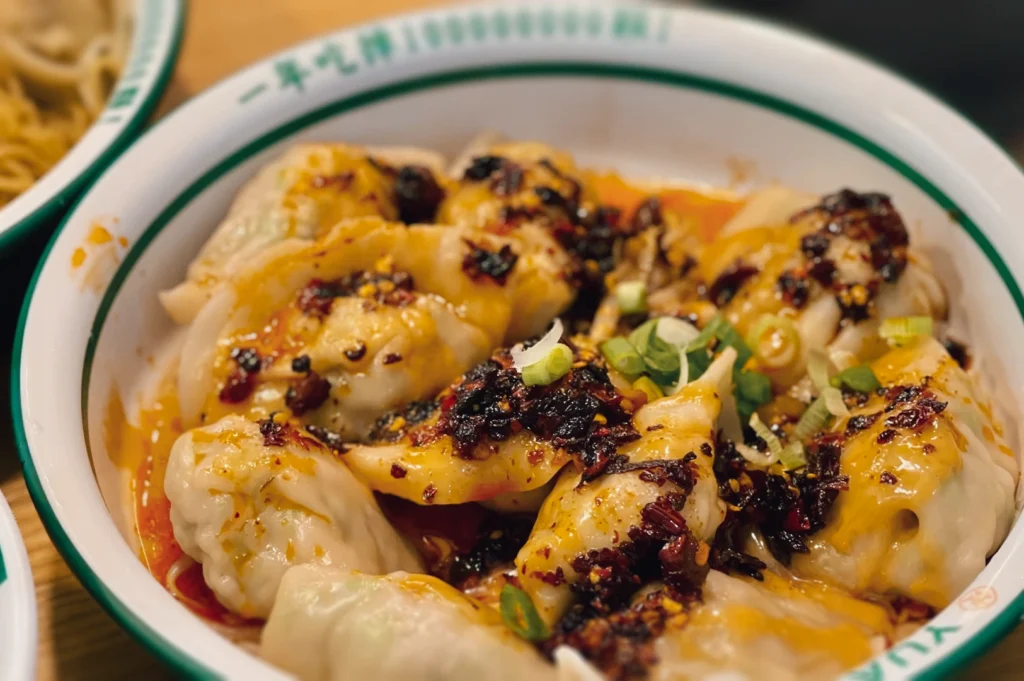 Macro close-up angled shot of juicy wontons drizzled with chili oil and peanut sauce showing texture and spice detail.