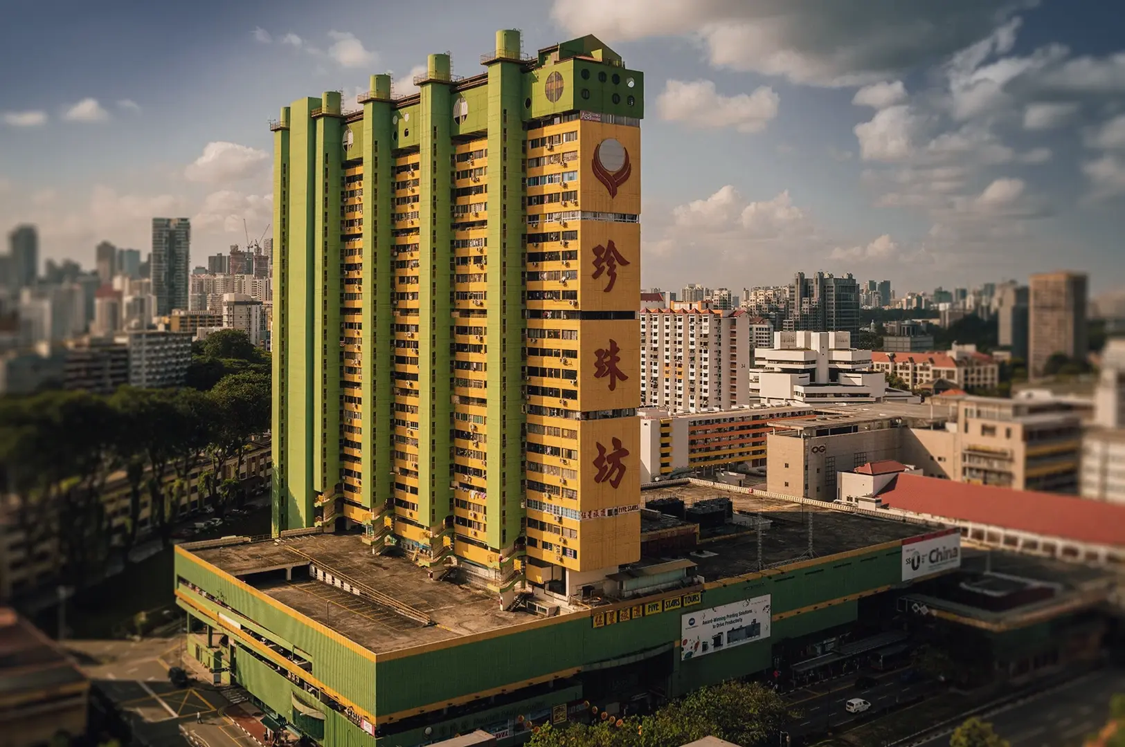 Low-angle wide shot of People’s Park Complex in Chinatown Singapore, showcasing green and yellow brutalist high-rise architecture above a podium shopping centre, surrounded by urban skyline and residential buildings under daylight