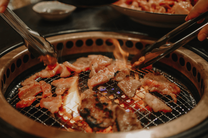 Close-up of sliced meat sizzling over open flames on a traditional Japanese circular charcoal grill, being actively turned by metal tongs.
