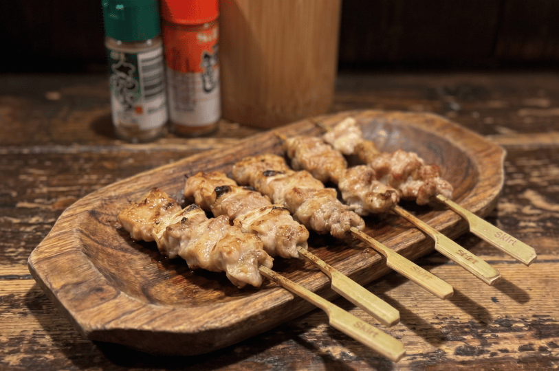 Six perfectly grilled yakitori chicken skewers served on a rustic carved wooden platter, with Japanese spice bottles in the soft-focus background.