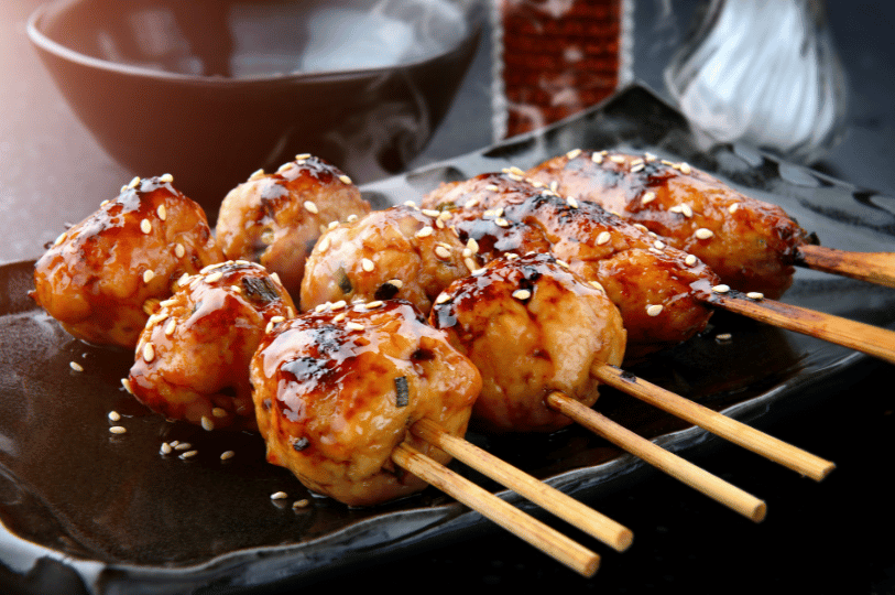 Close-up of glossy, sauce-glazed tsukune (Japanese chicken meatball) skewers sprinkled with white sesame seeds on a dark rectangular plate.