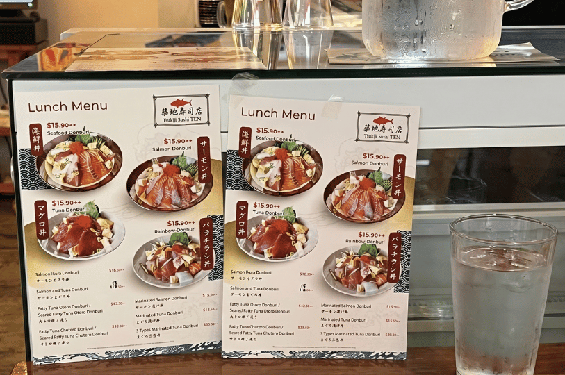 Tabletop lunch menus displaying various Japanese donburi seafood bowls, positioned next to a glass of ice water on a counter.