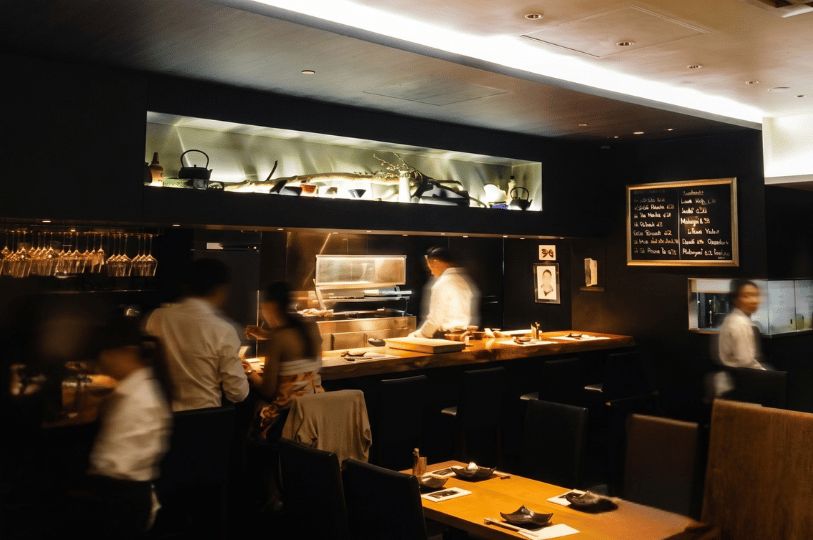 A warm, dimly lit interior of Kazu Sumiyaki featuring diners seated at a wooden counter while chefs prepare food in the open kitchen.