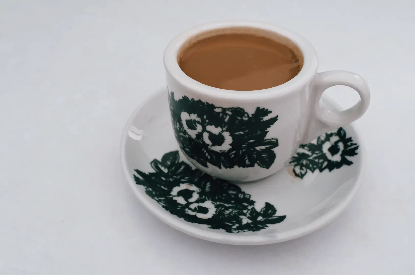 ose-up, slightly top-down shot of a white ceramic cup filled with light-brown traditional kopi, sitting on a matching saucer decorated with dark green floral motifs, set against a plain white background; soft, even lighting highlights the glossy ceramic surface, the smooth texture of the brewed coffee, and the simple presentation commonly associated with Singapore kopitiam coffee.