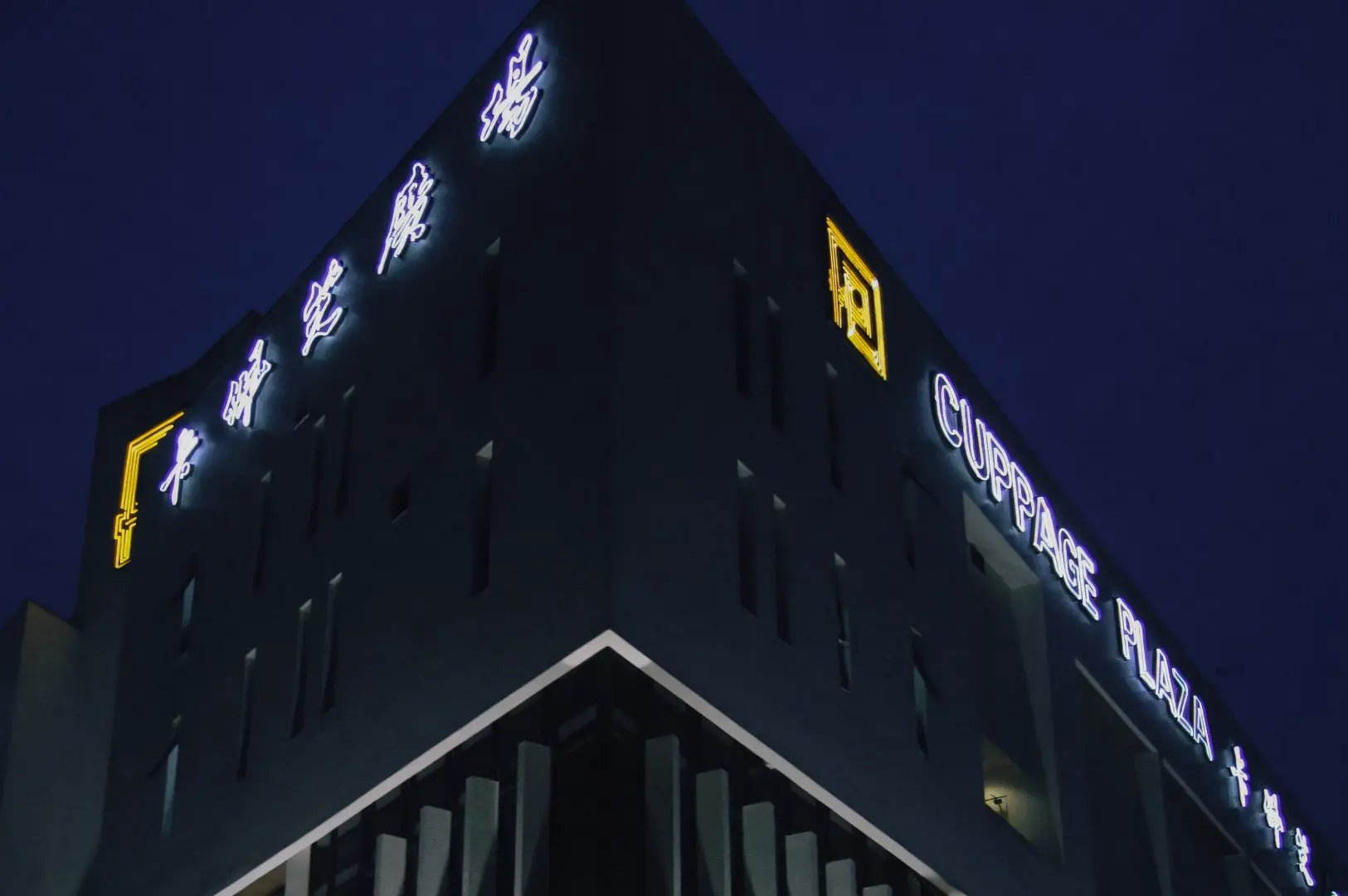Wide-angle, low-angle exterior shot of Cuppage Plaza at night, showing the dark, angular building facade with illuminated signage reading ‘Cuppage Plaza’ in English alongside glowing Japanese characters, set against a deep blue evening sky, with accent lighting highlighting the sharp geometry, concrete textures, and recessed windows of the structure.