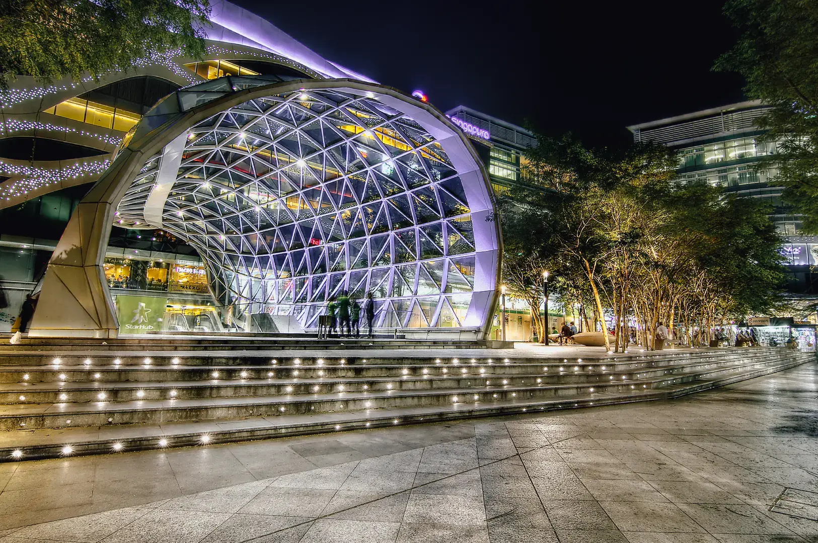Low-angle wide shot of Plaza Singapura’s modern glass dome entrance illuminated at night, showcasing iconic Orchard Road architecture and lively urban atmosphere in Singapore.