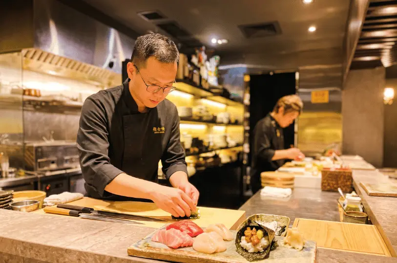 A focused sushi chef in a black uniform carefully prepares a dish behind a sushi bar, with a beautifully plated selection of fresh sashimi, scallops, and a hand roll in the foreground.