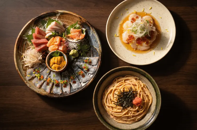 An elegant overhead view of a Japanese dinner spread on a dark wooden table, showcasing a large premium sashimi platter with sea urchin, a plate of scallop carpaccio garnished with salmon roe, and a bowl of creamy Mentaiko pasta topped with nori.