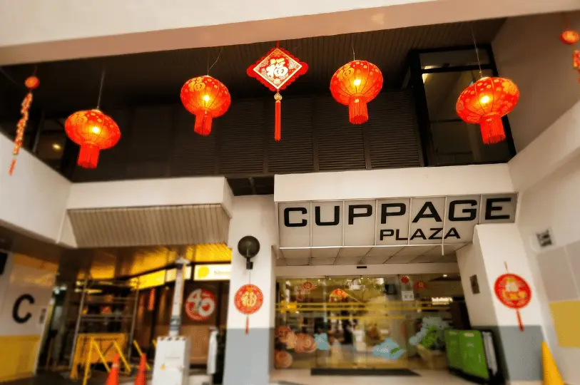 The exterior entrance of Cuppage Plaza in Singapore, featuring its prominent building signage and decorative red Chinese lanterns hanging above the glass doorways.