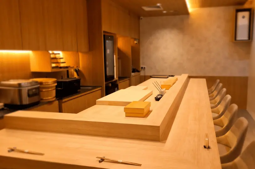 The serene, minimalist interior of a high-end Sushimasa Omakase, highlighting a pristine, light-wood L-shaped sushi counter lined with elegant matching chairs.