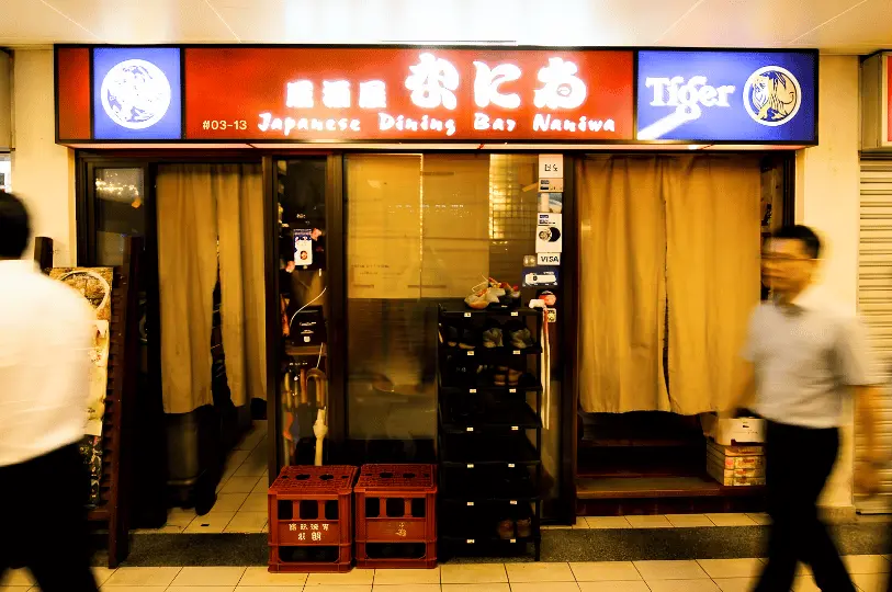izakaya-naniwa-sg- (1).webp: The unassuming storefront of Izakaya Naniwa featuring a red and blue illuminated sign, beige entrance curtains, and a small shoe rack, with blurred pedestrians walking past.