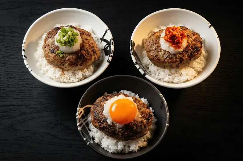 Three bowls of Japanese hamburg steak served over white rice with varying toppings, including a bright raw egg yolk, offering customizable options for different family members' tastes.