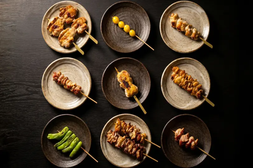 A grid of nine rustic ceramic plates, each displaying a different grilled yakitori skewer, including various chicken cuts, ginkgo nuts, and shishito peppers.