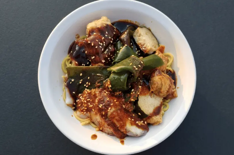 A close-up, top-down shot of a white bowl filled with vegetarian Hakka Yong Tau Foo over noodles, featuring stuffed tofu, mock meats, and green vegetables, all generously drizzled with a thick, dark sweet sauce and sprinkled with sesame seeds.