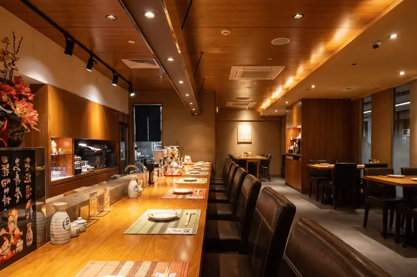 The warm and inviting interior of Hanashizuku Japanese Cuisine, featuring a long, brightly lit wooden counter with leather seating and cozy dining tables in the background.