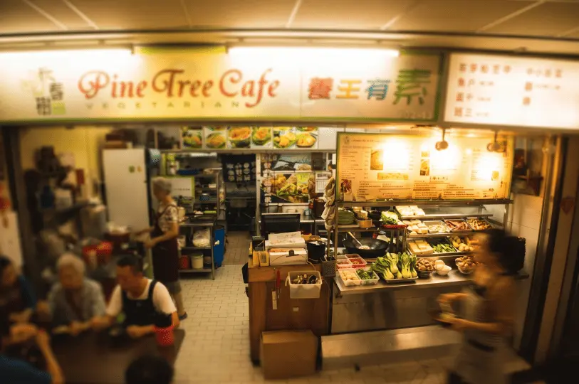 A bustling, slightly blurred wide shot of the Pine Tree Cafe stall, capturing the lively atmosphere with staff preparing food behind a counter stacked with fresh vegetables, and customers seated at tables on the left.
