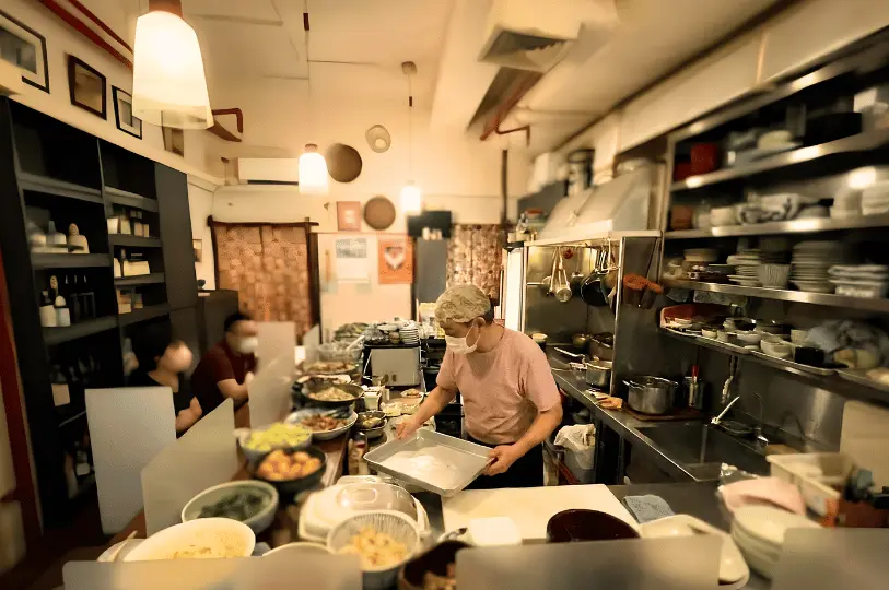 An older chef in a pink shirt and face mask works diligently behind the narrow counter of Izakaya Naniwa, with stainless steel kitchen equipment behind him and diners seated at the bar.