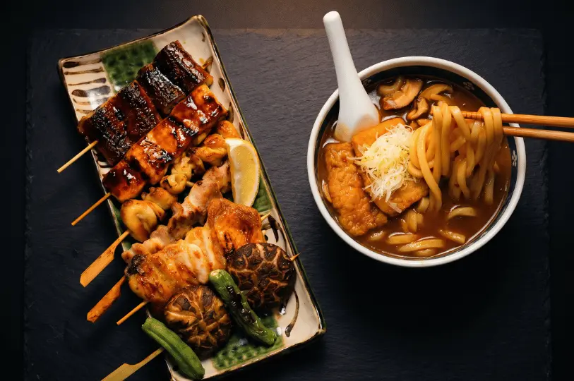 A top-down view of a Japanese meal on a dark slate background, featuring a rectangular plate of assorted yakitori skewers and a bowl of hearty curry udon noodles being lifted by chopsticks.