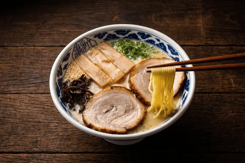 A steaming, rich bowl of Japanese tonkotsu ramen topped with tender chashu pork slices and bamboo shoots, with chopsticks lifting a portion of noodles.
