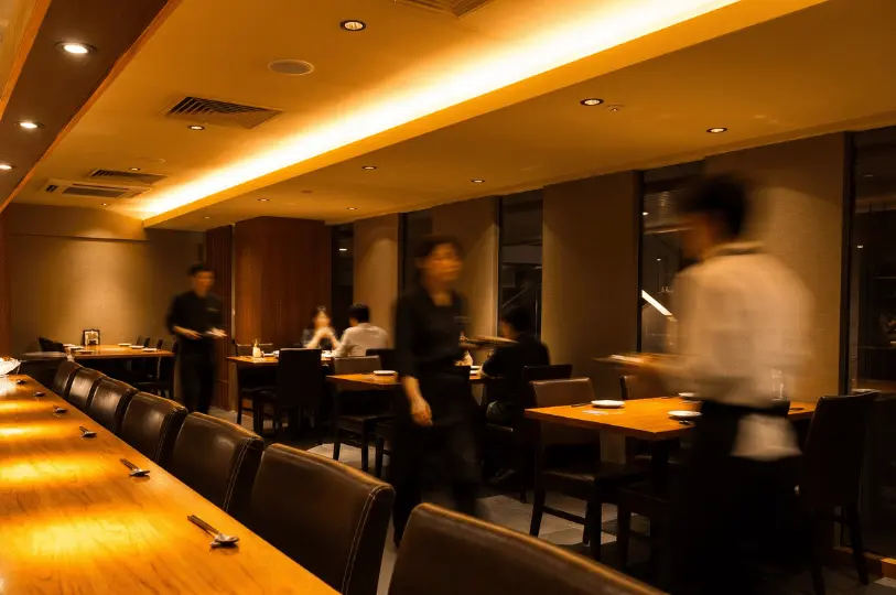 The softly lit dining room of Hanashizuku restaurant in motion, with the blurred movement of waitstaff briskly serving customers seated at intimate wooden tables.