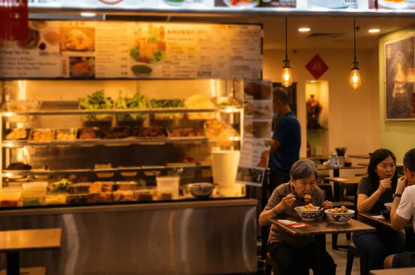 Diners enjoying comfort food inside the warmly lit Pine Tree Cafe, focusing on an elderly woman eating a bowl of noodles in the foreground, with the stall's food display case blurred in the background.