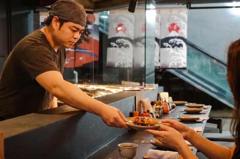 A chef at Kai Yakitori handing a freshly grilled chicken skewer on a plate directly to a customer across the dining counter.