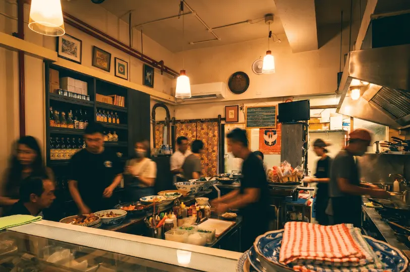 Inside the warmly lit Izakaya Naniwa, showing a bustling, cozy atmosphere with customers dining at the counter and staff moving around the open kitchen.