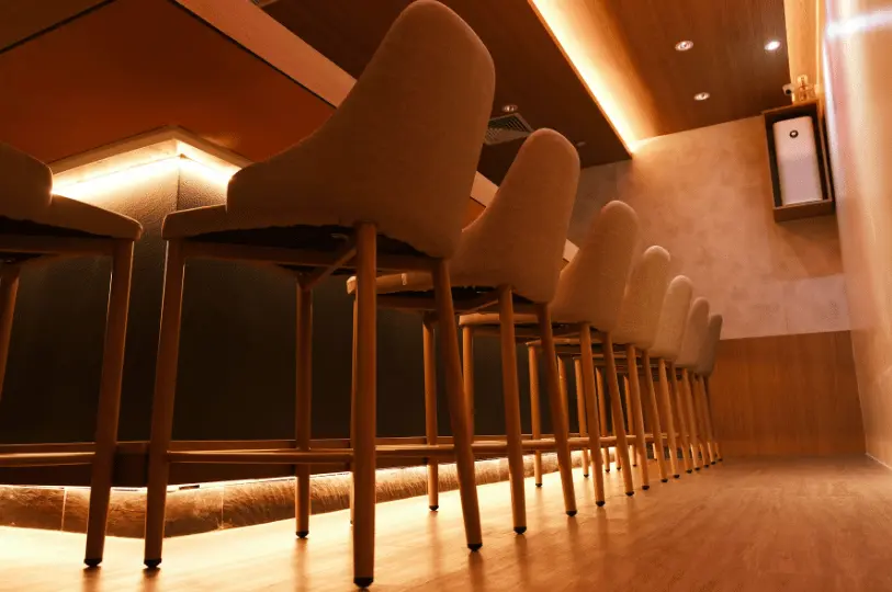 A low-angle shot of Sushimasa Omakase focusing on a neat row of wooden bar stools with tan fabric seating, lined up along a sushi counter featuring warm, under-counter LED lighting.