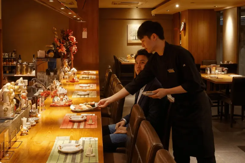 A chef courteously serving a fresh plate of sushi to a dining patron seated at the wooden counter of Hanashizuku, which is adorned with traditional Japanese lucky cat figurines.