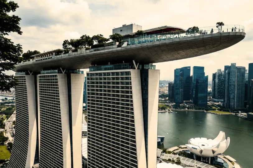 An aerial view of the iconic Marina Bay Sands hotel in Singapore, showing its three towers, the boat-shaped rooftop sky deck, and the lotus-shaped Art-Science Museum below.