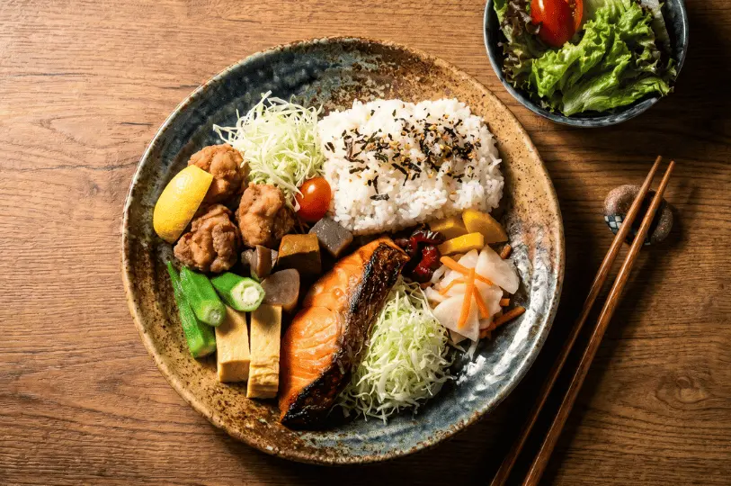 A hearty and colorful Japanese meal served on a rustic ceramic plate, featuring a portion of grilled salmon, crispy chicken karaage, rolled omelet, rice topped with furikake, a side salad, and an assortment of fresh vegetables and pickles.