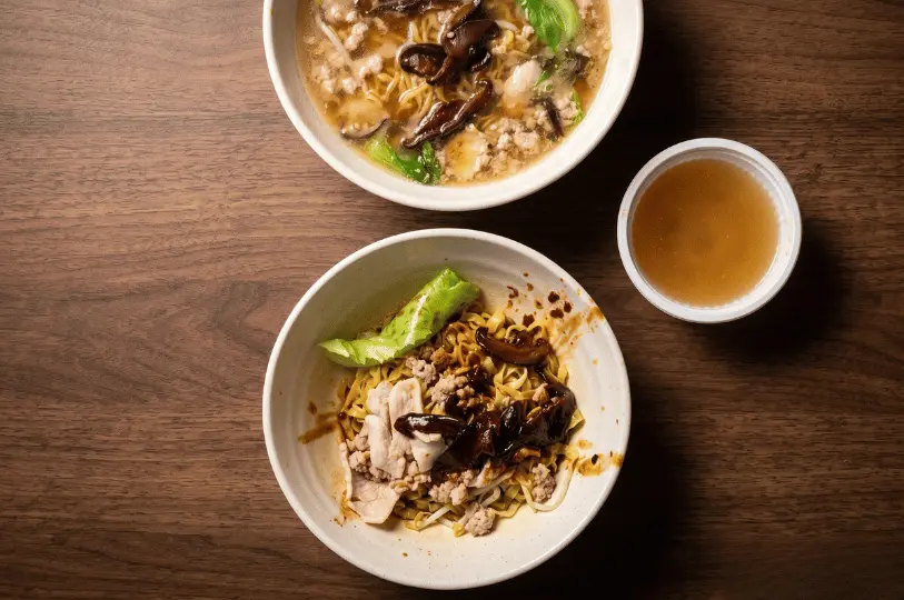 Two bowls of vegetarian noodles on a wooden table: the top bowl contains a warm noodle soup with mushrooms and greens, while the bottom bowl features dry noodles mixed with dark sauce, sliced mushrooms, and mock minced meat, served with a small side of clear broth.