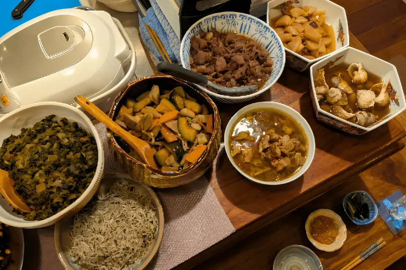 A top-down view of several large bowls filled with homestyle Japanese obanzai dishes, including stewed meats, root vegetables, and simmered greens, resting on a wooden counter.