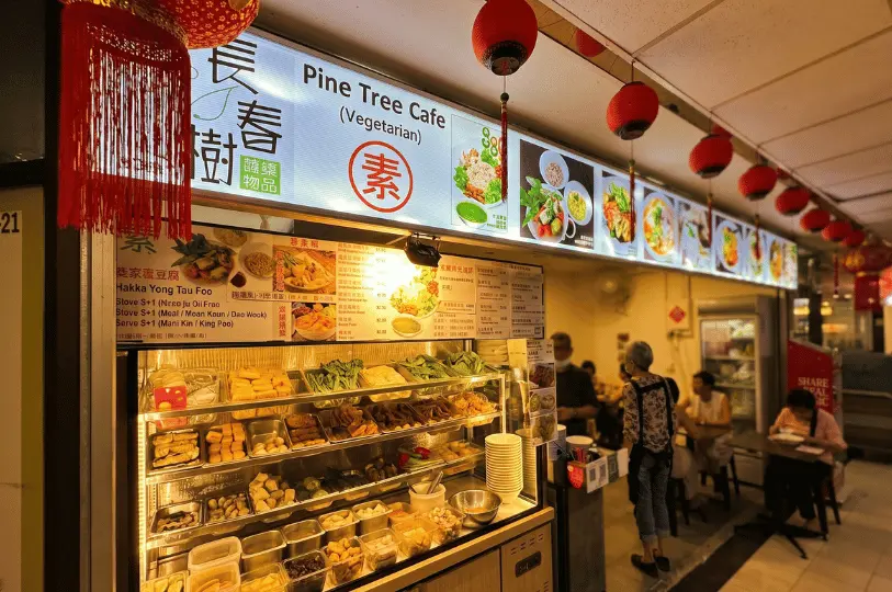 The brightly lit storefront of Pine Tree Cafe in Fortune Centre, featuring a display case packed with fresh vegetarian ingredients like tofu and greens, overhead menus, hanging red lanterns, and customers dining at nearby tables.