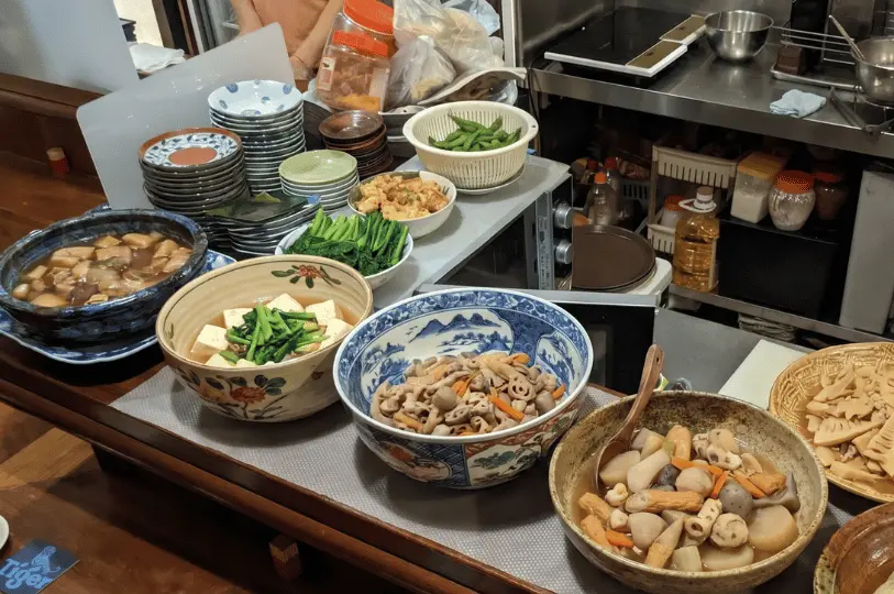 A close-up of the counter display showing an array of freshly prepared Japanese comfort foods, such as stewed tofu, lotus root, and fishcakes, in large decorative ceramic bowls.